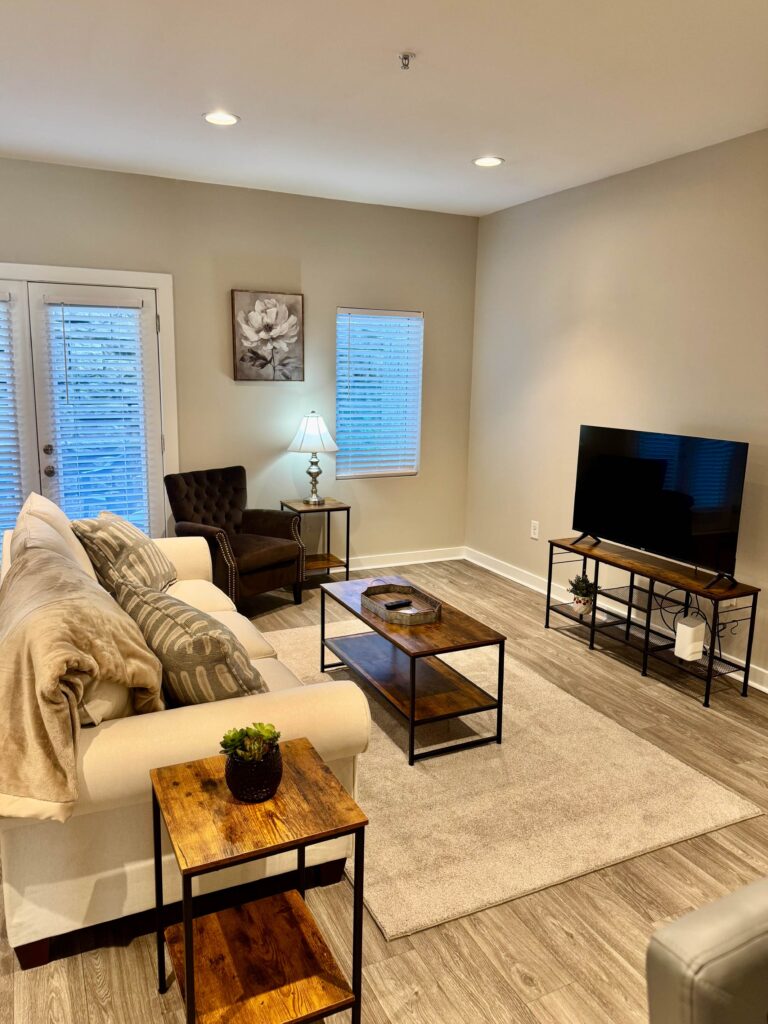 Comfortable living room featuring a large sofa, accent chair, flat-screen television, coffee table, and private patio access in a bright open-concept apartment.