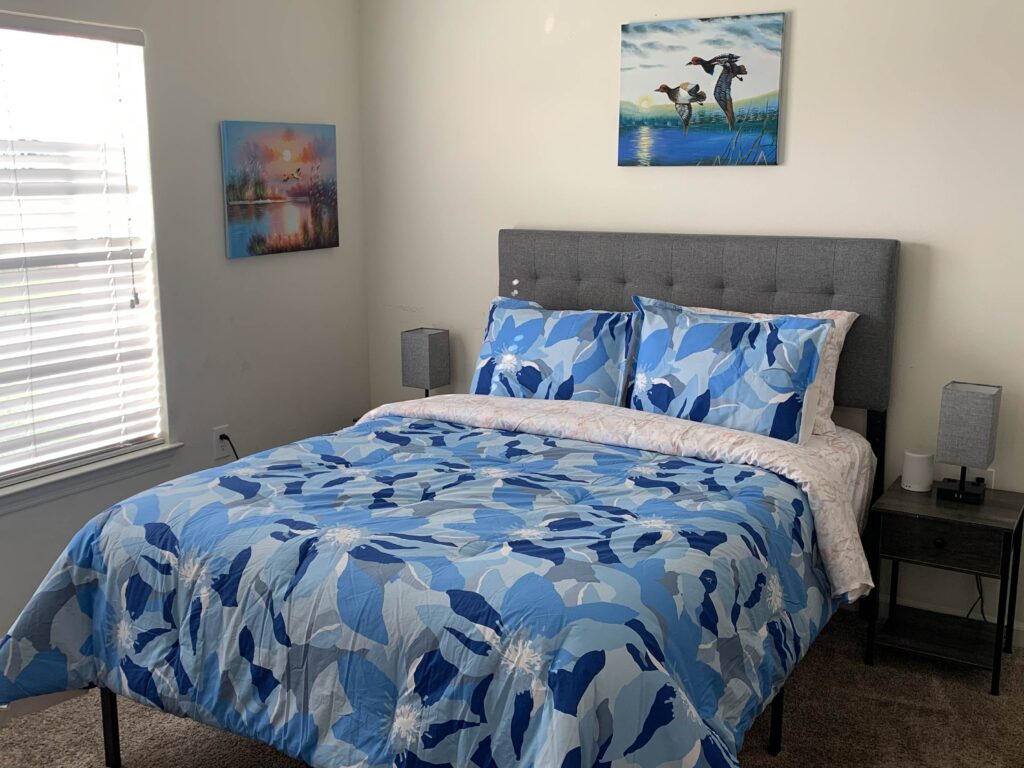 Bedroom with blue floral bedding, gray upholstered headboard, bedside tables, and framed artwork.