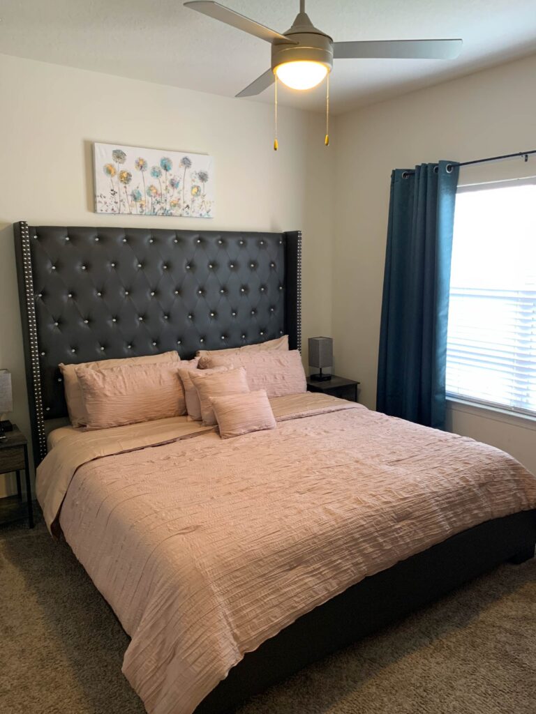 Bedroom with tufted upholstered headboard, neutral bedding, bedside tables, and blackout curtains.