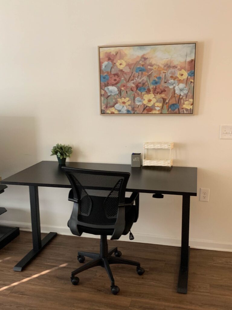 Minimal workspace with black desk, rolling office chair, floral wall art, and hardwood flooring.