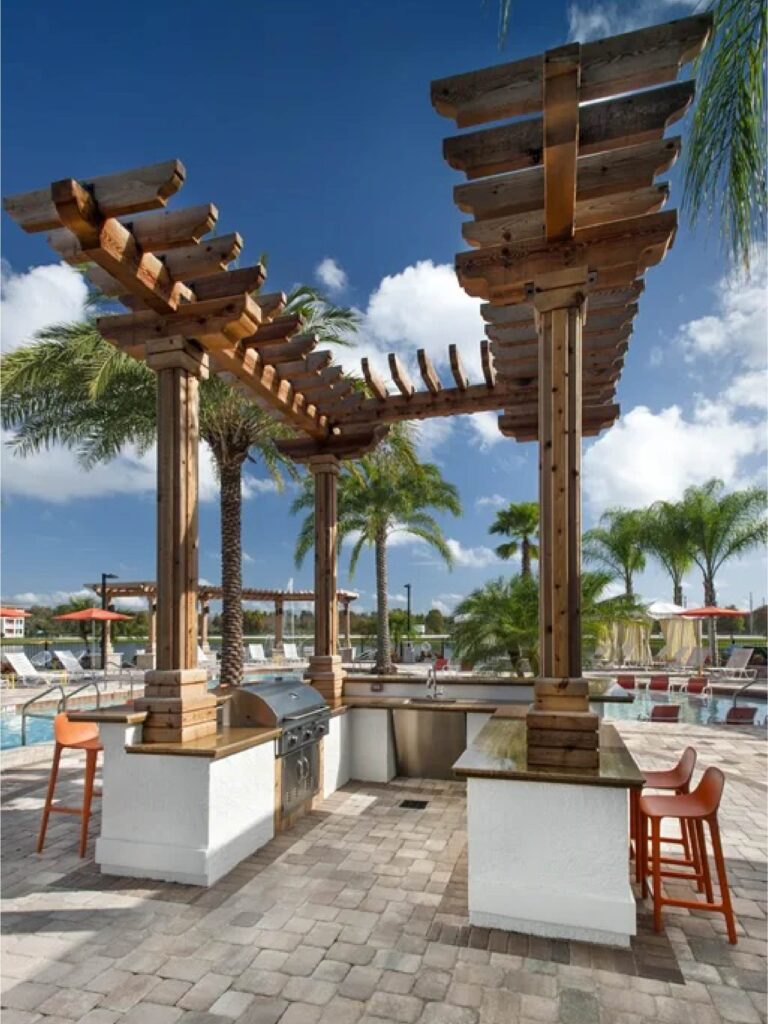 Outdoor resort style pool area with pergola covered grilling station, palm trees, lounge seating, and sunny skies.