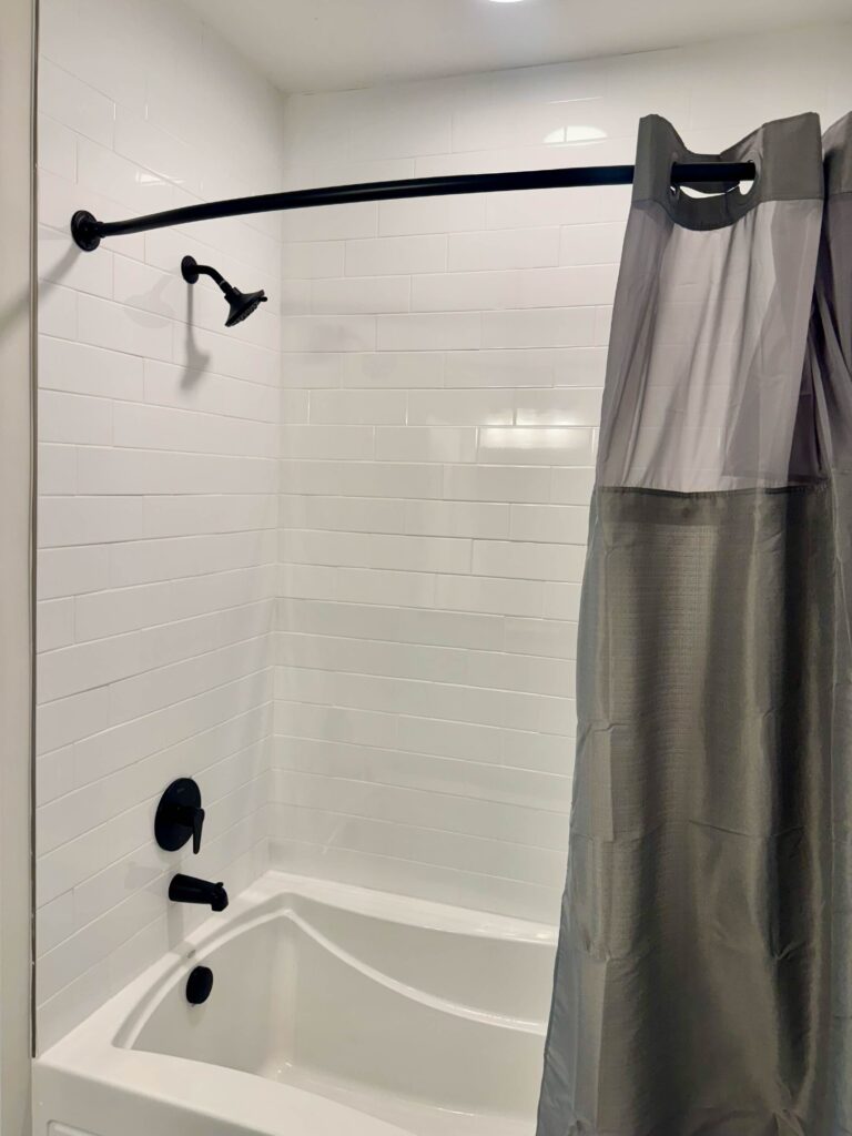 Modern guest bathroom featuring a full-size bathtub and shower combination with white subway tile surround, matte black fixtures, and a curved shower curtain rod for additional space.
