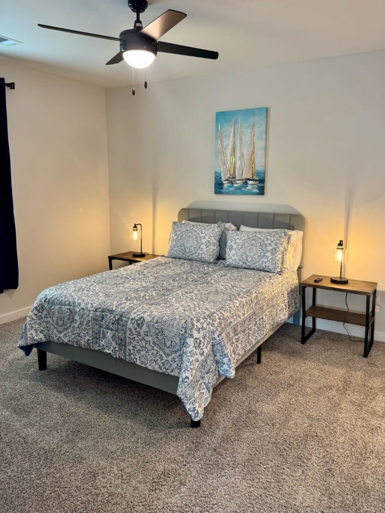 Comfortably furnished bedroom featuring a queen-size bed, soft carpeting, modern nightstands, bedside lighting, ceiling fan, and coastal-inspired artwork.