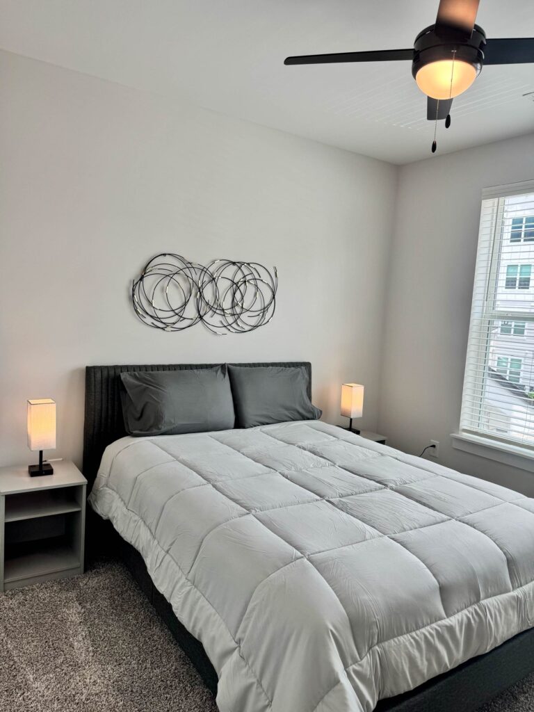 Modern guest bedroom featuring a queen-size bed with gray bedding, contemporary nightstands, accent lighting, ceiling fan, plush carpeting, and decorative wall art.
