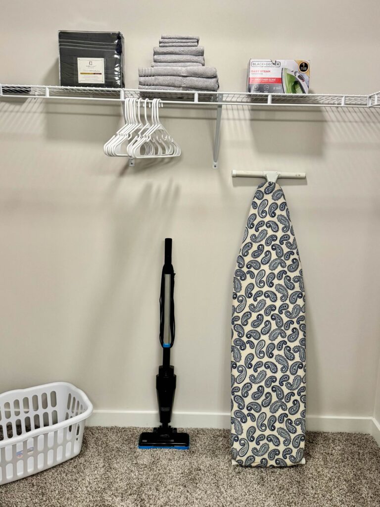 Spacious walk-in closet featuring hanging space, storage shelving, extra linens, towels, clothes hangers, an ironing board, steam iron, laundry basket, and cordless vacuum.