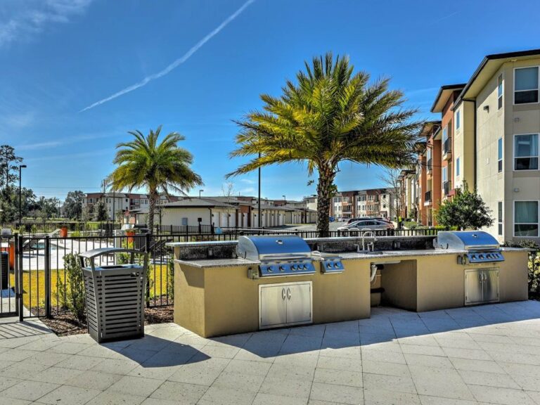 Community outdoor grilling area at Maxwell Apartments featuring multiple stainless steel gas grills, food preparation space, palm trees, and resort style landscaping.