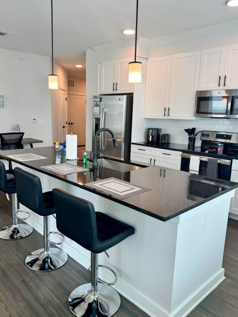 Modern gourmet kitchen at Maxwell Apartments featuring white shaker cabinetry, stainless steel appliances, granite countertops, oversized island seating, and pendant lighting.