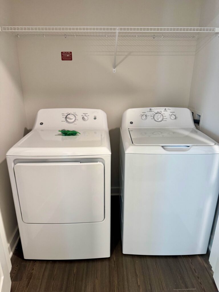 Private in-unit washer and dryer at Maxwell Apartments featuring full-size laundry appliances and convenient storage shelving.