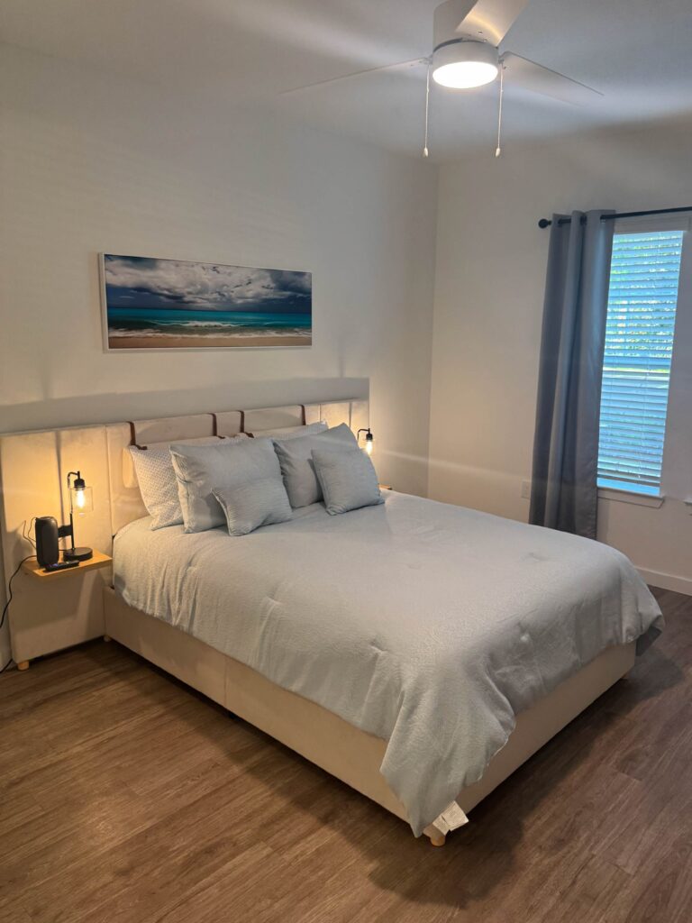 Modern bedroom with upholstered king bed, soft neutral decor, wood-style flooring, and bedside lighting.