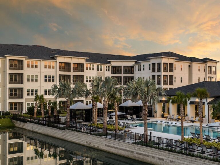Resort style pool and clubhouse area at sunset with palm trees, cabanas, waterfront views, and modern apartment buildings.
