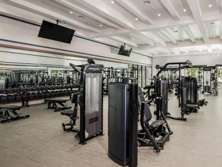 Modern resident fitness center with strength machines, free weights, benches, mirrored walls, and mounted televisions.
