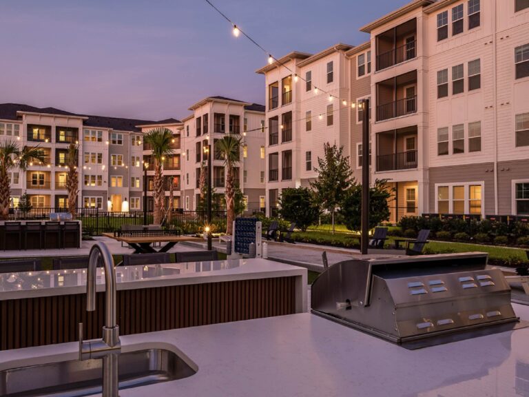 Outdoor grilling and entertainment area with stainless steel grills, string lights, seating areas, and modern apartment buildings at dusk.