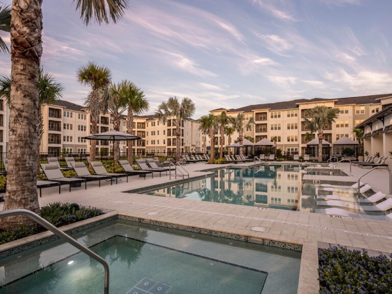Resort-style swimming pool with spa area, palm trees, lounge seating, and modern apartment buildings at sunset.