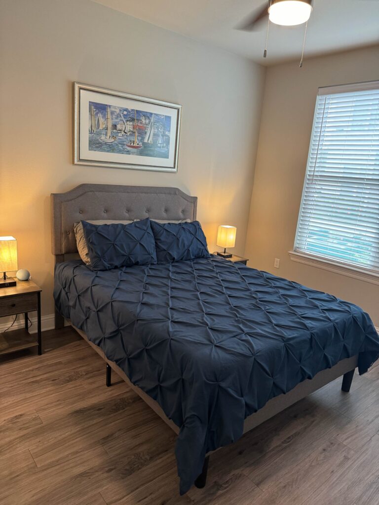 Comfortable bedroom featuring a queen bed with upholstered headboard, bedside tables, warm lighting, ceiling fan, and large window with natural light.