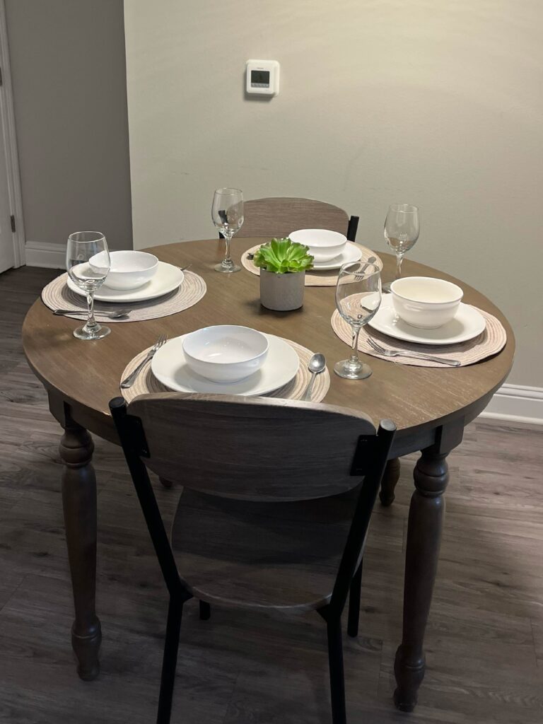 Round dining table set for four with place settings, stemware, placemats, and modern seating in a comfortable dining area.
