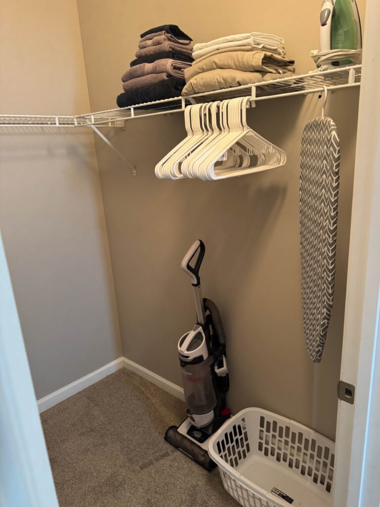 Walk in closet with folded towels, hangers, ironing board, vacuum, and guest storage space.