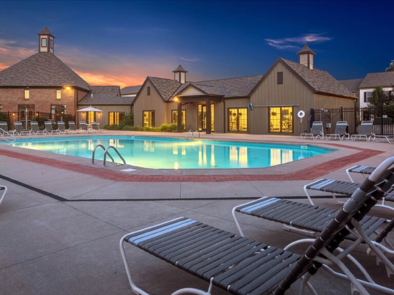 Resort style swimming pool at dusk with lounge chairs, illuminated clubhouse buildings, and relaxing outdoor seating areas.