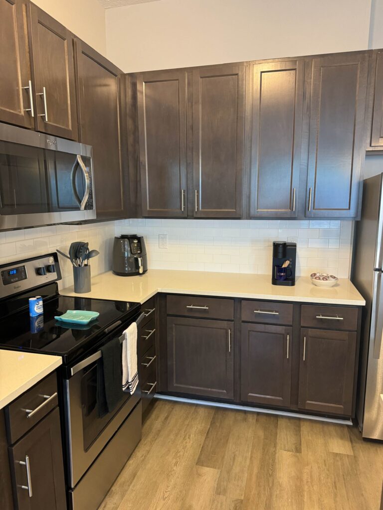 Modern kitchen at Tapestry Bocage featuring dark wood cabinetry, stainless steel appliances, quartz countertops, subway tile backsplash, coffee maker, and air fryer.