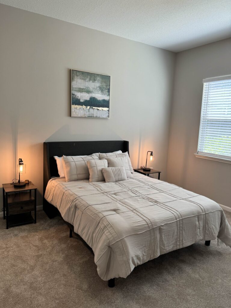 Primary bedroom at Tapestry Bocage featuring a queen bed with neutral bedding, bedside tables with lamps, plush carpeting, and a large window providing natural light.