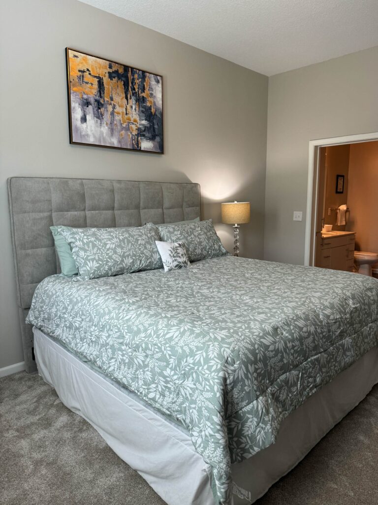 Primary bedroom at Tapestry Bocage featuring a king bed with patterned bedding, upholstered headboard, bedside lamp, plush carpeting, and an attached en-suite bathroom.