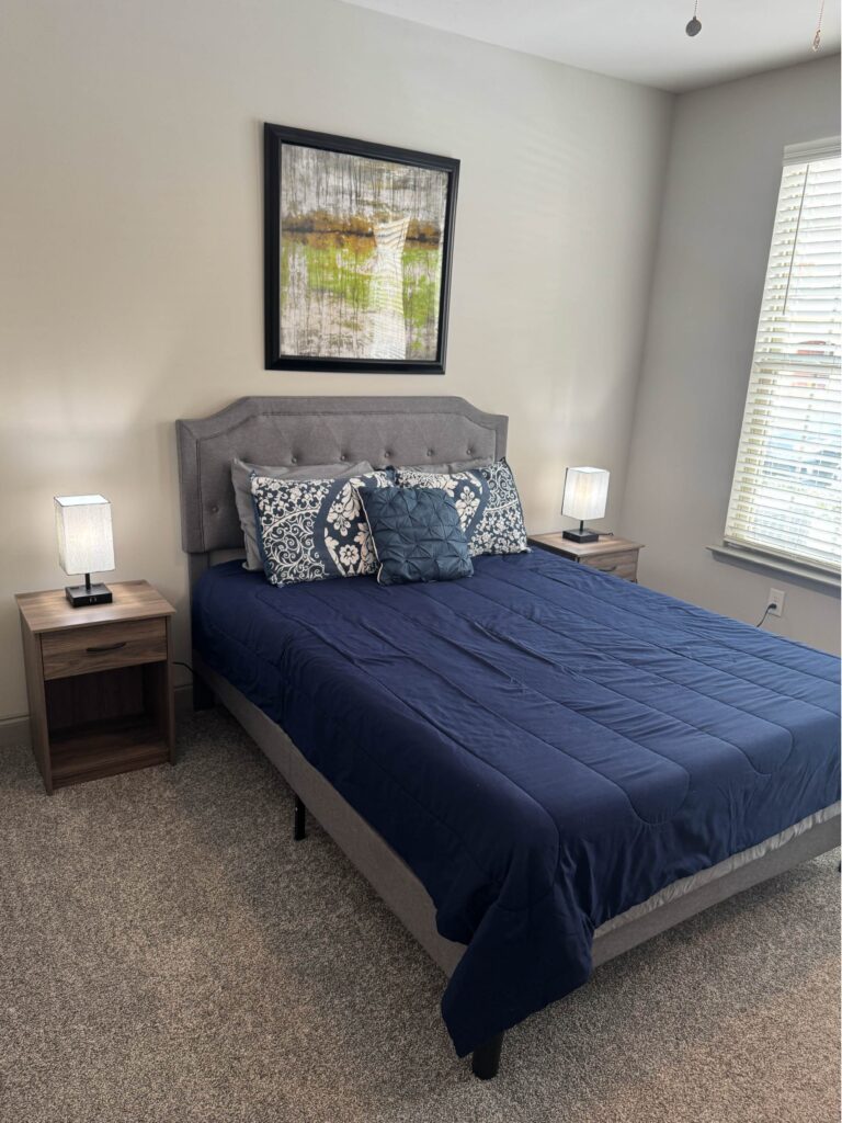 Furnished bedroom with blue bedding, upholstered headboard, bedside lamps, carpeted flooring, and modern wall art at The Hub in Bowling Green, Kentucky.