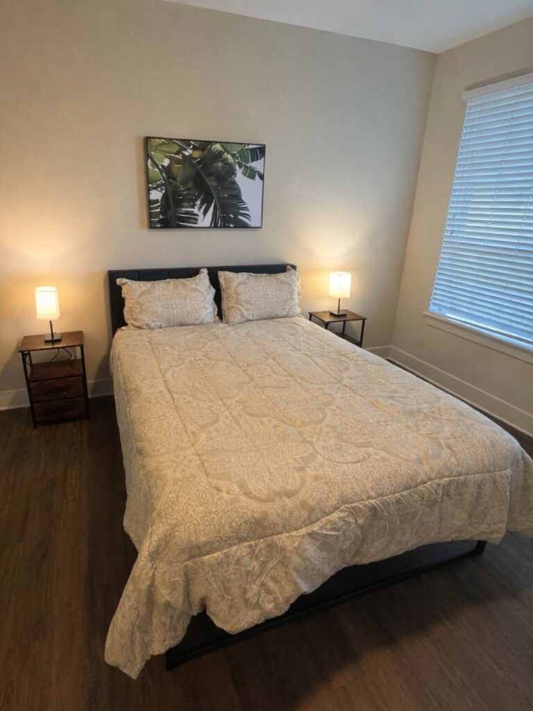 Minimalist furnished bedroom with a queen bed, matching bedside lamps, wood flooring, and tropical wall art.