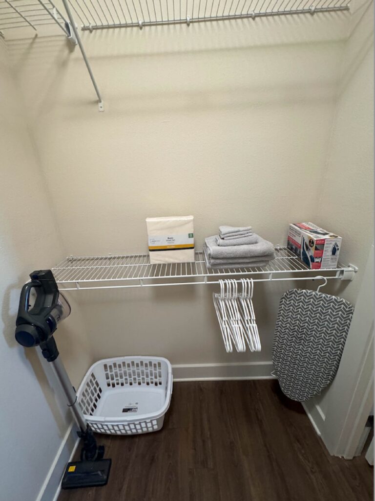 Walk in closet with shelving, ironing board, extra linens, vacuum, and storage space.