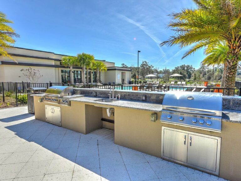 Outdoor grilling and entertainment area with stainless steel BBQ grills, pool views, and palm trees.