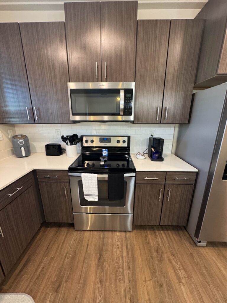 Modern apartment kitchen with stainless steel appliances, dark wood cabinetry, white countertops, and wood-style flooring.