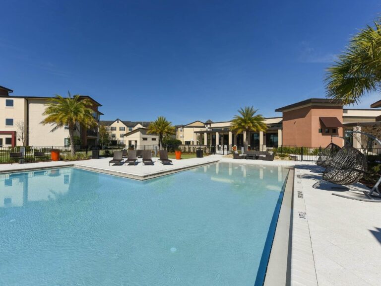 Resort-style swimming pool with lounge seating, palm trees, and modern apartment buildings in the background.