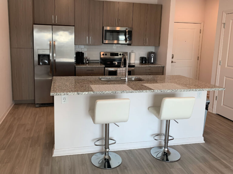 Modern open-concept kitchen at V2 Apartments in Fort Myers with granite island seating, stainless steel appliances, and contemporary wood cabinetry.