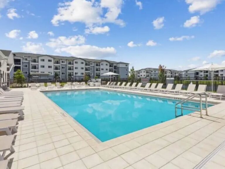 Resort style swimming pool at V2 Apartments in Fort Myers with white lounge chairs, modern apartment buildings, and expansive sun deck under a bright blue sky.