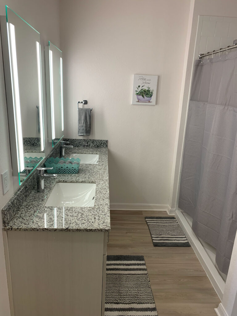 Modern bathroom at V2 Apartments in Fort Myers featuring dual granite vanities, illuminated mirrors, and a walk in shower with contemporary finishes.
