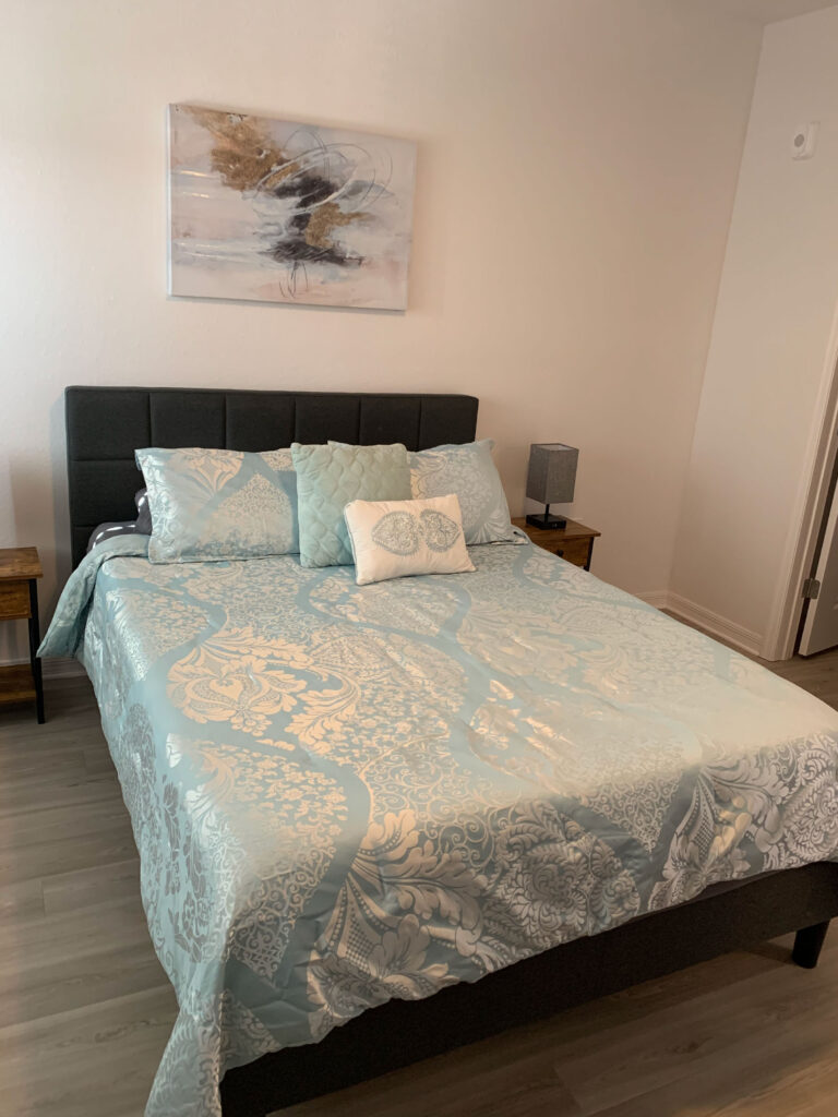 Modern furnished bedroom at V2 Apartments in Fort Myers featuring a queen bed, decorative bedding, bedside tables, and contemporary wall art.