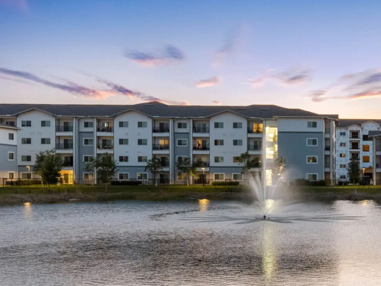 Waterfront exterior view of V2 Apartments in Fort Myers at sunset featuring modern apartment buildings overlooking a pond with illuminated fountain.