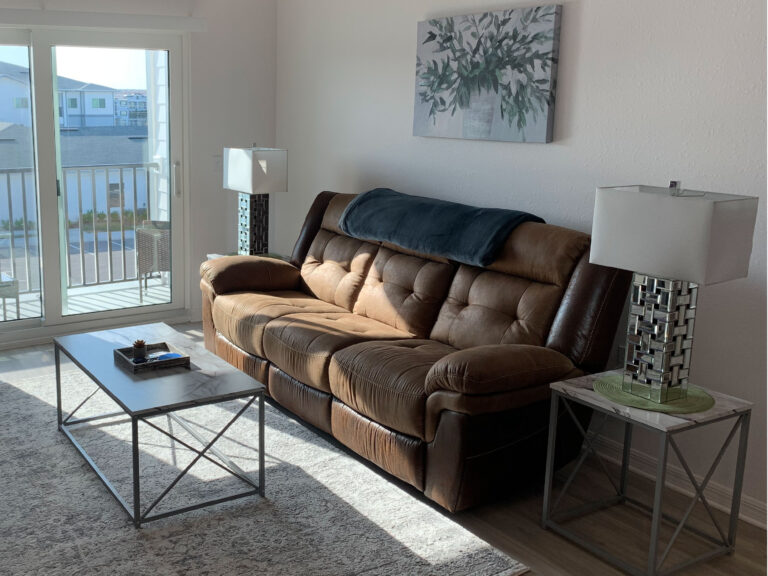 Modern furnished living room at V2 Apartments in Fort Myers featuring a brown reclining sofa, contemporary décor, coffee table, and private balcony access.
