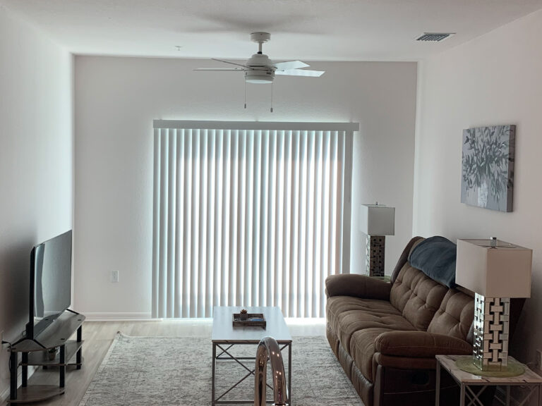 Modern living room at V2 Apartments in Fort Myers with leather reclining sofa, large TV, ceiling fan, and floor-to-ceiling sliding glass doors.