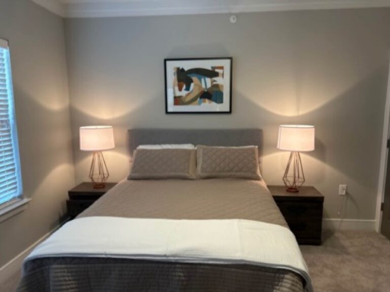 Primary bedroom at Vale Apartments featuring a comfortable queen-size bed, bedside tables with lamps, plush carpeting, modern artwork, and a relaxing residential atmosphere.