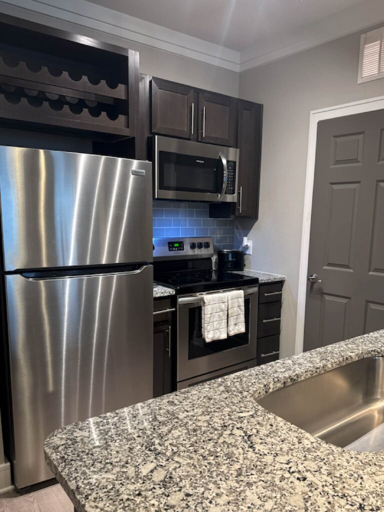 Modern kitchen at Vale Apartments featuring stainless steel appliances, granite countertops, dark wood cabinetry, tile backsplash, built-in microwave, and a spacious breakfast bar.