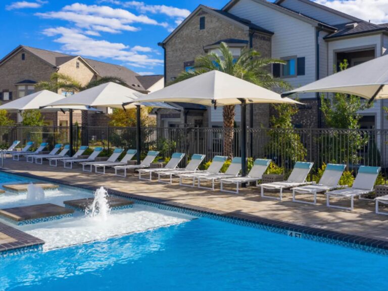 Resort-style swimming pool at Vale Apartments in the Spring/Houston area featuring expansive sundecks, shaded lounge seating, water features, palm trees, and upscale apartment homes.