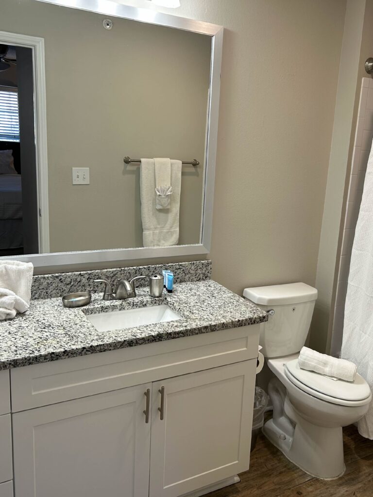 Modern full bathroom featuring a granite vanity countertop, white cabinetry, large framed mirror, bathtub and shower combination, and fresh white towels.