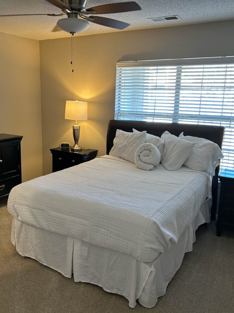 Spacious primary bedroom at Vista Commons in Columbia, South Carolina, featuring a queen-size bed, nightstands, dresser, ceiling fan, and large window with natural light.