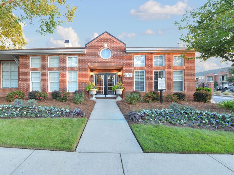 Exterior view of the Vista Commons clubhouse in Columbia, South Carolina, featuring classic brick architecture, landscaped grounds, colorful flower beds, and a welcoming community entrance.