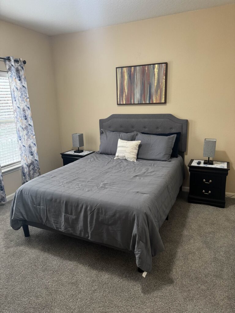 Spacious primary bedroom featuring a queen-size bed, upholstered headboard, matching nightstands, bedside lamps, and large windows providing natural light at Waterstone at Brier Creek in Lexington, Kentucky.