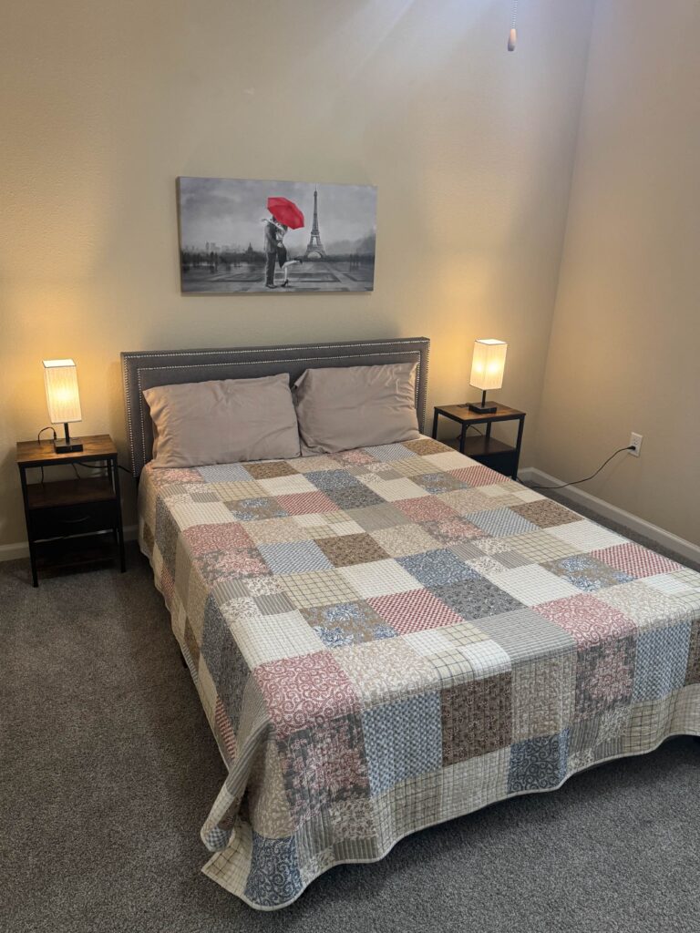 Secondary bedroom at Waterstone at Brier Creek featuring a queen bed with patchwork quilt bedding, upholstered headboard, matching bedside tables with lamps, soft carpeting, and wall art depicting the Eiffel Tower.