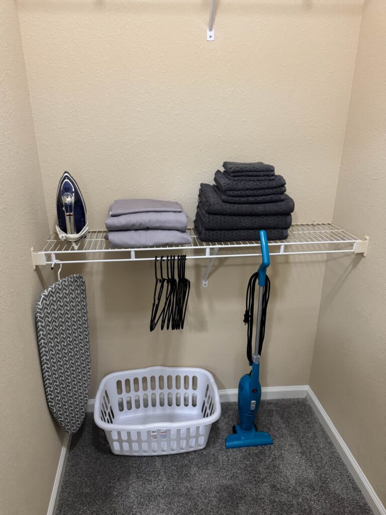 Walk-in closet at Waterstone at Brier Creek featuring hanging space, fresh linens, towels, hangers, ironing equipment, laundry basket, and cleaning tools for guest convenience.