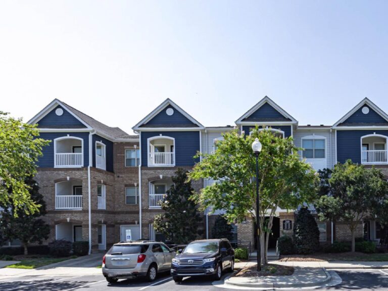 Exterior view of Waterstone at Brier Creek featuring three-story residential buildings with brick and blue siding accents, private balconies, mature landscaping, and convenient resident parking.