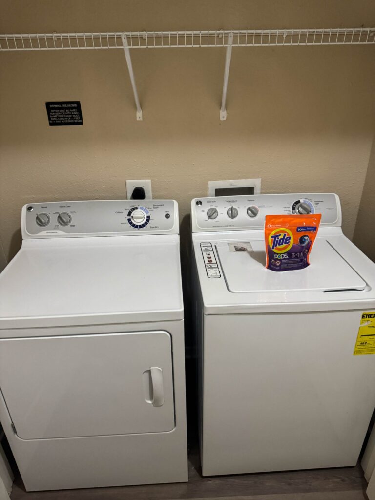 In-unit laundry area featuring a full-size washer and dryer with overhead storage shelving and complimentary laundry supplies at Waterstone at Brier Creek in Lexington, Kentucky.