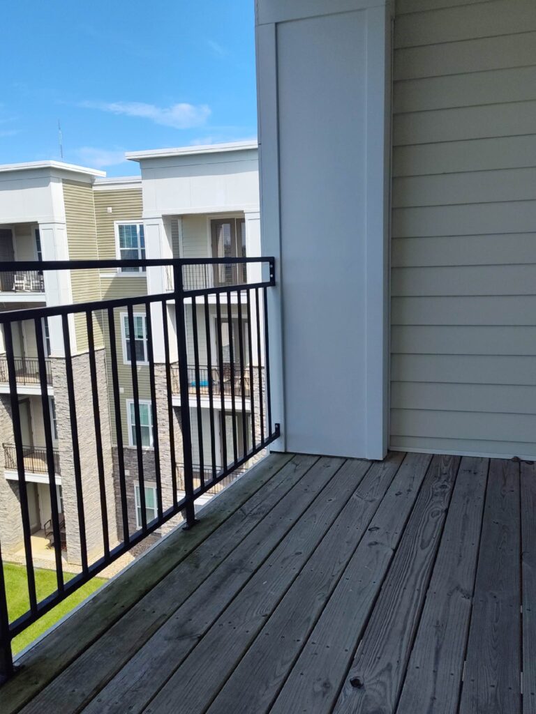 Private apartment balcony with wood-style flooring, black railing, and views of neighboring residential buildings.
