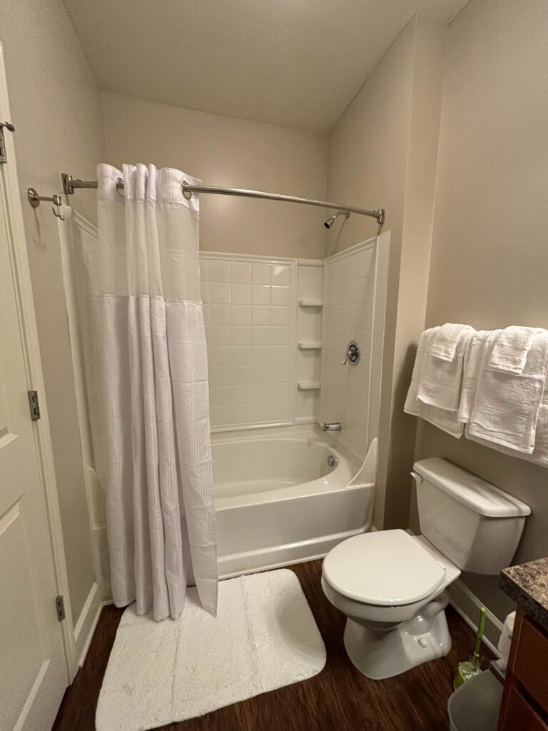 Full bathroom featuring a tub and shower combination with white tile surround, white shower curtain, toilet, fresh towels, and wood-style flooring.